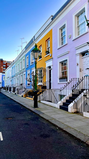 A row of vibrantly painted terraced houses in Kensington, with colors ranging from pastel blue to lavender, featuring white-framed sash windows and black wrought-iron railings. A lamppost is positioned on the sidewalk, which is paved with concrete slabs. The properties have small steps leading up to their front doors, some with potted plants or decorative elements in window boxes. The image is taken during daylight, with a clear blue sky overhead. This scene illustrates a typical residential street in Kensington suitable for house removals, where professional relocation services by Removal Companies Kensington might operate, involving moving furniture, packing materials, and equipment to facilitate home relocation and furniture transport.