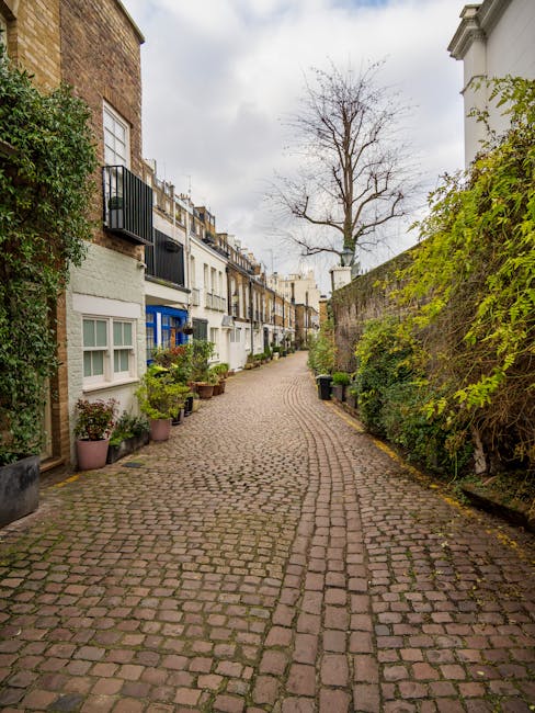 A narrow cobblestone street in Kensington with residential buildings on the left side, featuring white and pastel-colored facades, some with small balconies and potted plants, while the right side is bordered by a high brick wall covered with green climbing plants. In the background, a leafless tree stands against an overcast sky. The street appears quiet with no visible vehicles or pedestrians, illustrating a typical housing area suitable for home relocation or furniture transport services provided by companies like Removal Companies Kensington. The scene highlights the quaint, historic character of the neighborhood, emphasizing the environment where professional removals can assist with packing, loading, and transport during house removals or moving logistics.