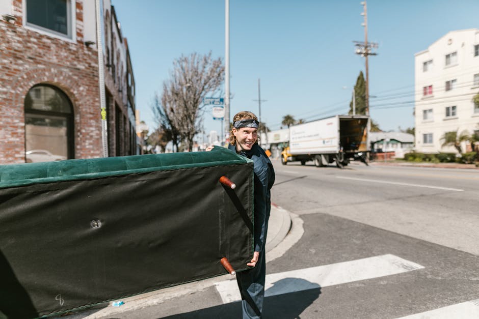 A young man with a cheerful expression, wearing a black jacket and a headband, is outdoors on a city street near Gloucester Road, engaged in a home relocation activity. He is carrying a large, black moving blanket with green padding, which is partially folded and secured with metal handles, indicating furniture protection and handling during the packing and moving process. The background features a brick building, leafless trees, and a busy street with a white delivery truck and cars, suggesting a typical urban environment near Earls Court in Kensington. The scene takes place on the pavement adjacent to a curb, under natural daylight with clear skies. This image illustrates the logistics involved in furniture transport, part of a professional removals service by Removal Companies Kensington, supporting the preparation and execution of house or flat moves in the Kensington area, including packing, loading, and transportation as part of comprehensive moving services.