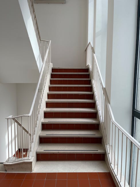 A set of indoor stairs with white metal railings, leading upwards within a building on the left and right sides. The stairs are made of brown tiles with a white edge, and the steps are clearly visible from the ground floor landing. To the right of the staircase, there is a large window allowing natural light to illuminate the area, showcasing the neutral-colored walls and modern interior. The space appears clean and well-maintained, suitable for a home relocation or moving process. Removal Companies Kensington have implemented these stairs to facilitate the transport of furniture and belongings between floors, highlighting the importance of careful handling and logistical planning during house removals in the W14 area and Kensington.