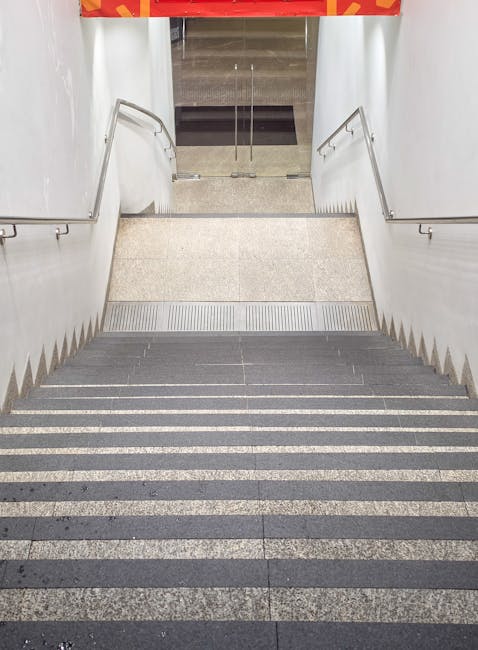 A downward view of a staircase leading to a basement or lower level, with dark grey and light grey non-slip treads and metal handrails on both sides. At the bottom of the stairs, there is a small landing area with a textured metal strip across the doorway, which appears to be an entrance to an elevator or corridor. The surrounding walls are white, and natural light illuminates the internal space. This staircase is part of a building in Kensington used by Removal Companies Kensington for home relocation and furniture transport, illustrating the challenging stair access they often manage during removals in the W14 area.