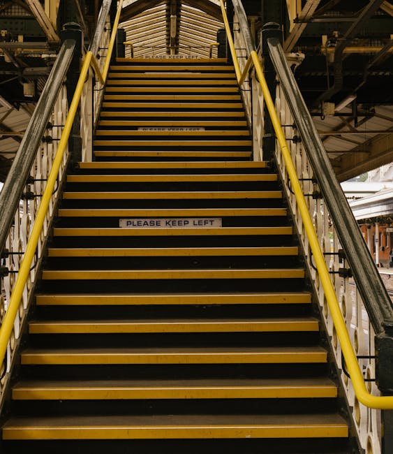 A set of indoor stairs with white metal railings, leading upwards within a building on the left and right sides. The stairs are made of brown tiles with a white edge, and the steps are clearly visible from the ground floor landing. To the right of the staircase, there is a large window allowing natural light to illuminate the area, showcasing the neutral-colored walls and modern interior. The space appears clean and well-maintained, suitable for a home relocation or moving process. Removal Companies Kensington have implemented these stairs to facilitate the transport of furniture and belongings between floors, highlighting the importance of careful handling and logistical planning during house removals in the W14 area and Kensington.