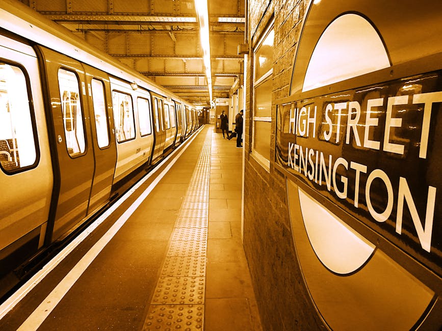 Inside the underground station at High Street Kensington, a train is stationary along the platform, with its doors closed. The platform surface features tactile paving strips for accessibility, running parallel to the train. Several people are visible in the distance, some standing near the platform edge, observing safety guidelines during the home relocation or moving process. The station interior is illuminated with warm lighting, and a large roundel sign displaying 'HIGH STREET KENSINGTON' is mounted on a brick wall to the right. The setting includes station features such as a wooden wall panel, glass display cases, and metal fixtures, indicating a typical London Underground environment. This image reflects the environment where furniture transport or packing activities by [COMPANY_NAME], a professional removals company in Kensington, might occur, especially during moving logistics involving station transfers or transportation of household belongings.