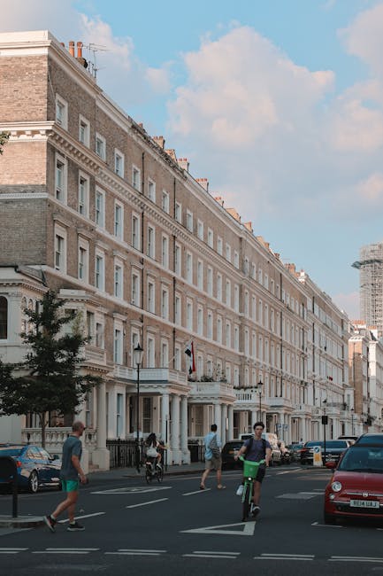A street scene in Kensington showing a row of elegant, multi-storey Victorian terraced houses with cream and beige facades, decorative cornices, and large sash windows. Several people are seen on the pavement, including a cyclist riding along the road, pedestrians walking, and a person carrying a cardboard box, indicating a home relocation process. Parked cars, including a red hatchback, line the curb, while a black van with its rear doors open is visible, suggesting loading or unloading activities typical of a professional removal service. The sky is partly cloudy with blue patches, and the environment appears well-lit by natural daylight. This scene reflects the typical logistics involved in removals and furniture transport within Kensington, where strict parking permits and local regulations often influence the moving process. The image illustrates the importance of adhering to local council rules for removals parking permits, supported by [COMPANY_NAME], specialists in removals and house moves in the area.