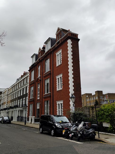 A multi-storey red brick building with white window frames and decorative white corner quoins stands prominently on a street lined with similar Victorian-style terraced houses. The building features dormer windows on the top floor under a pitched roof. In the foreground, a black van is parked on the pavement next to a cluster of scooters and motorbikes, likely involved in a home relocation or delivery process. The overcast sky and street lighting suggest daytime with diffused natural light. The environment appears residential, with a mix of buildings extending along the street, and some greenery visible in a small garden area behind a stone wall on the right. The scene reflects an urban setting suitable for house removals, with the vehicles and surroundings indicating ongoing moving or furniture transport operations, possibly coordinated by Removal Companies Kensington for a Kensington High Street property with narrow access.