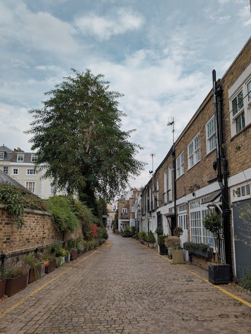 A row of Victorian terraced houses in Kensington W8 featuring pastel-colored facades with white window frames and decorative architectural details. The houses are set along a narrow paved pavement wit