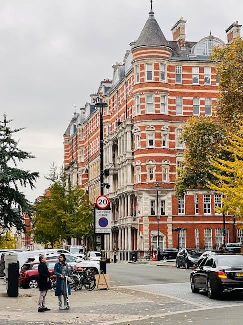 A street scene in South Kensington featuring a large, historic red-brick building with architectural details such as bay windows, decorative stonework, and a turreted corner tower. In the foreground, two individuals are engaged in conversation near parked bicycles and a black trash bin, with additional vehicles including cars parked along the street and driving past. Nearby, a road sign indicates a 20 mph speed limit and a no-parking zone. The area is bordered by trees with autumn foliage, and the street is populated with various parked vehicles, including a white van and a black sedan. This setting illustrates typical urban surroundings where house removals and furniture transport services provided by Removal Companies Kensington might operate, especially during home relocation or moving logistics involving street access and parking considerations.