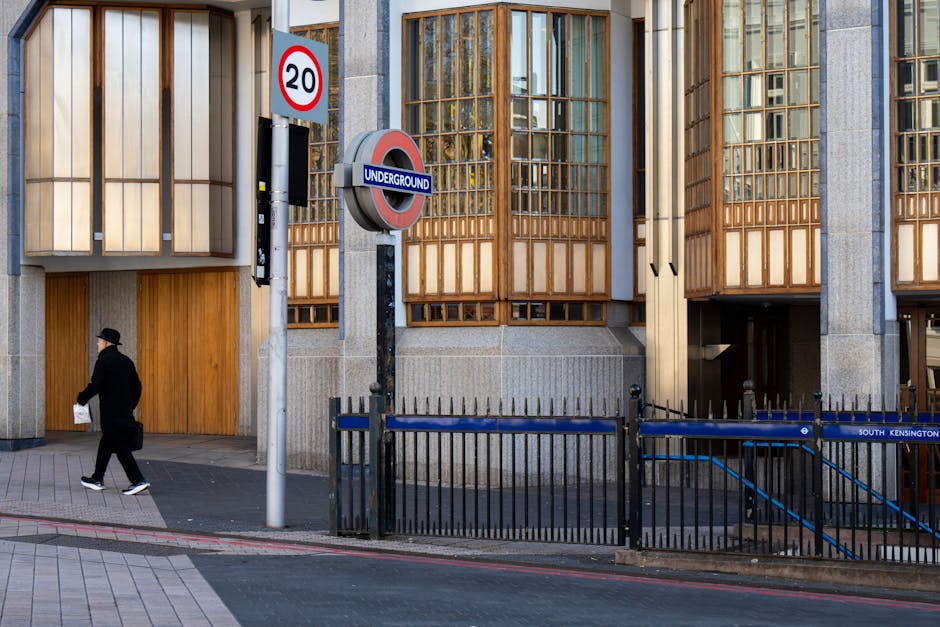Outside a modern building with wooden-framed windows and concrete walls, a black metal railing surrounds an underground entrance to South Kensington station, marked with a blue and red 'Underground' sign. A street sign indicates a 20 mph speed limit. On the sidewalk, a person dressed in dark clothing, including a coat and hat, is walking past holding a paper or document in one hand. The area appears to be a busy urban environment, possibly part of a home relocation or furniture transport process nearby, as the building's entrance could be accessed during removals or moving services. The scene is well-lit with natural daylight, and the street surface is paved with grey tiles and asphalt, with a slight curb separating the sidewalk from the road.