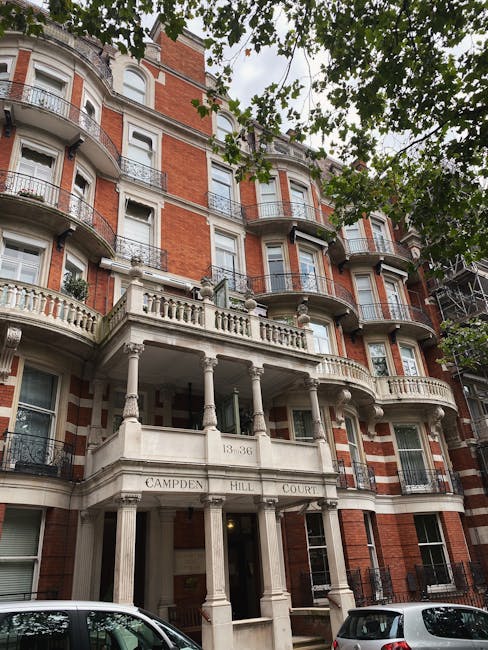 A multi-storey red brick building with ornate white stone balconies and decorative railings, situated on Campden Hill Court in South Kensington. The building features tall, arched windows and a prominent entrance supported by classical columns, with the address number 13-36 visible above the doorway. Green foliage from trees partially obscures the upper floors, and parked cars are visible along the street in front of the building. This image relates to house removals and the logistics of furniture transport, highlighting the challenges of moving large items through building exteriors, which can be affected by delays and elevator access issues during residential relocations.
